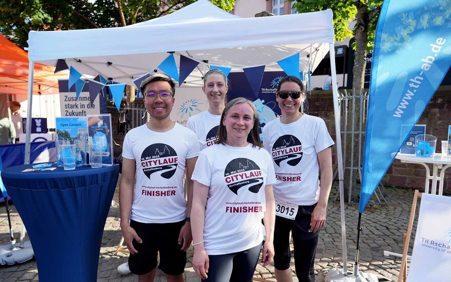 4 TH-Teilnehmende mit weißen Citylauf-Finisher-T-Shirts vor Stand der TH auf Schlossplatz