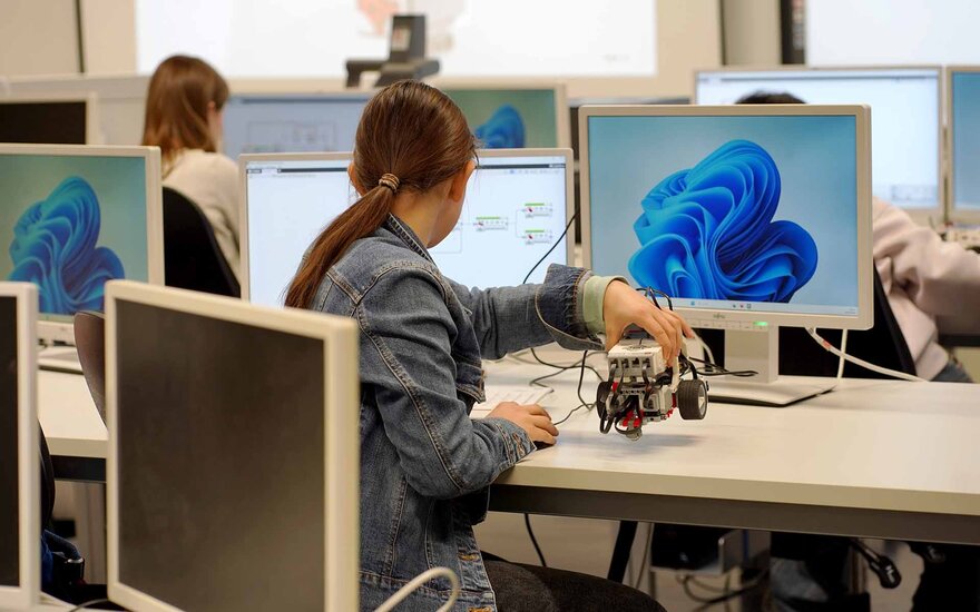 Ein Mädchen schaut sitzend auf einen Monitor und hält ein Legofahrzeug in der Hand.