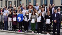 Gruppenbild mit rund 40 Personen, die teilweise Zertifikate halten, auf Treppenstufen vor den Neubauten