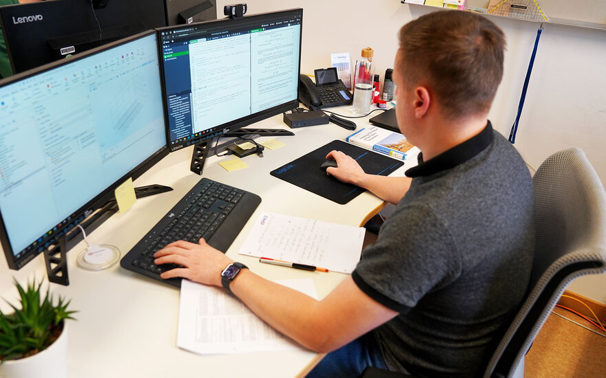 Luca Spielmann sitzt mit dem Rücken zur Kamera gewandt an seinem Arbeitsplatz, an dem zwei Monitore und ein Notizblock zu sehen sind.