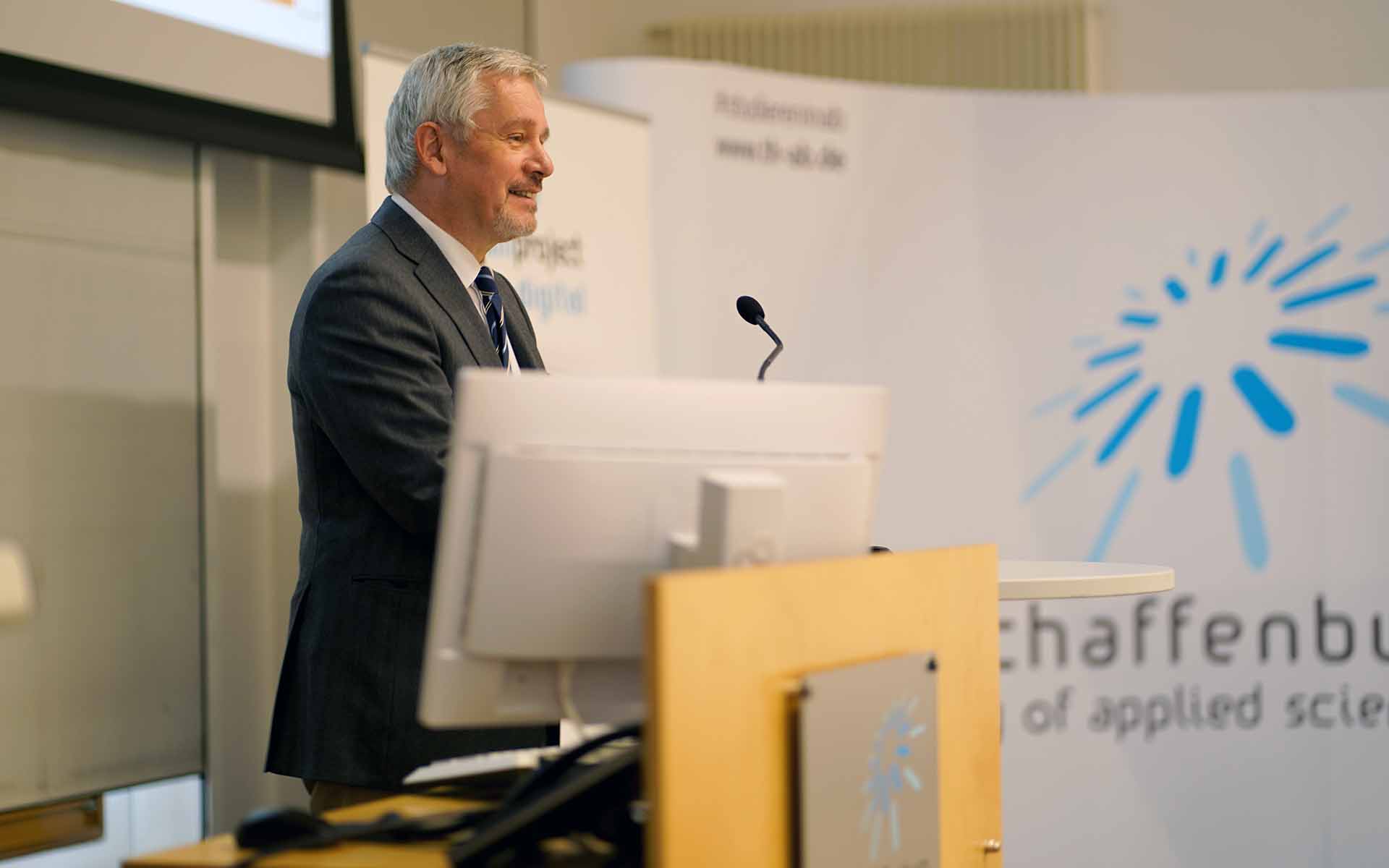 Professor Hofmann steht hinter einem Monitor am Rednerpult und spricht zum Publikum. Im Hintergrund ist eine Wand mit dem Logo der TH Aschaffenburg zu sehen.