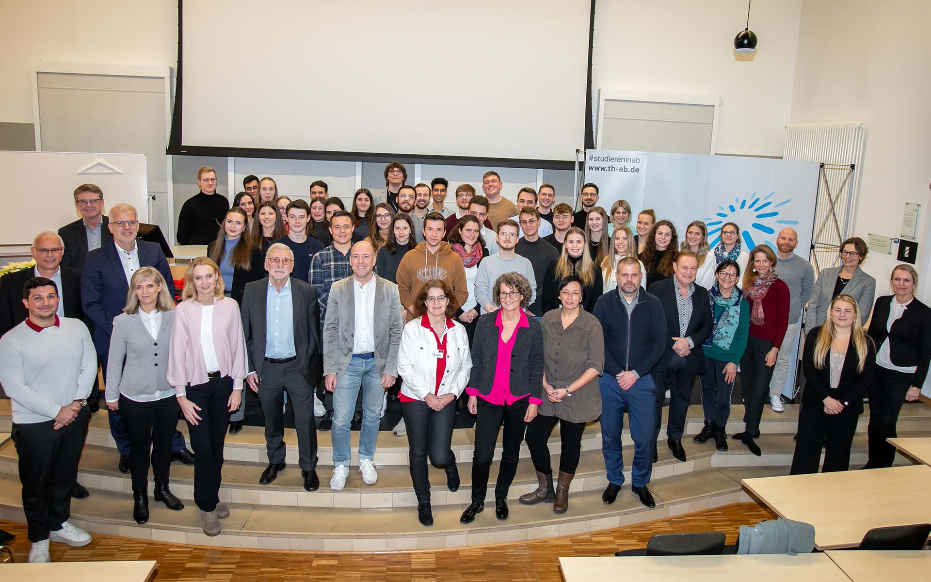 Fördernde, Stipendiatinnen und Stipendiaten stehen in der Aula der Hochschule in mehreren Reihen nebeneinander auf der Bühne, im Hintergrund ist ein Teil des blauen TH-Logos zu sehen
