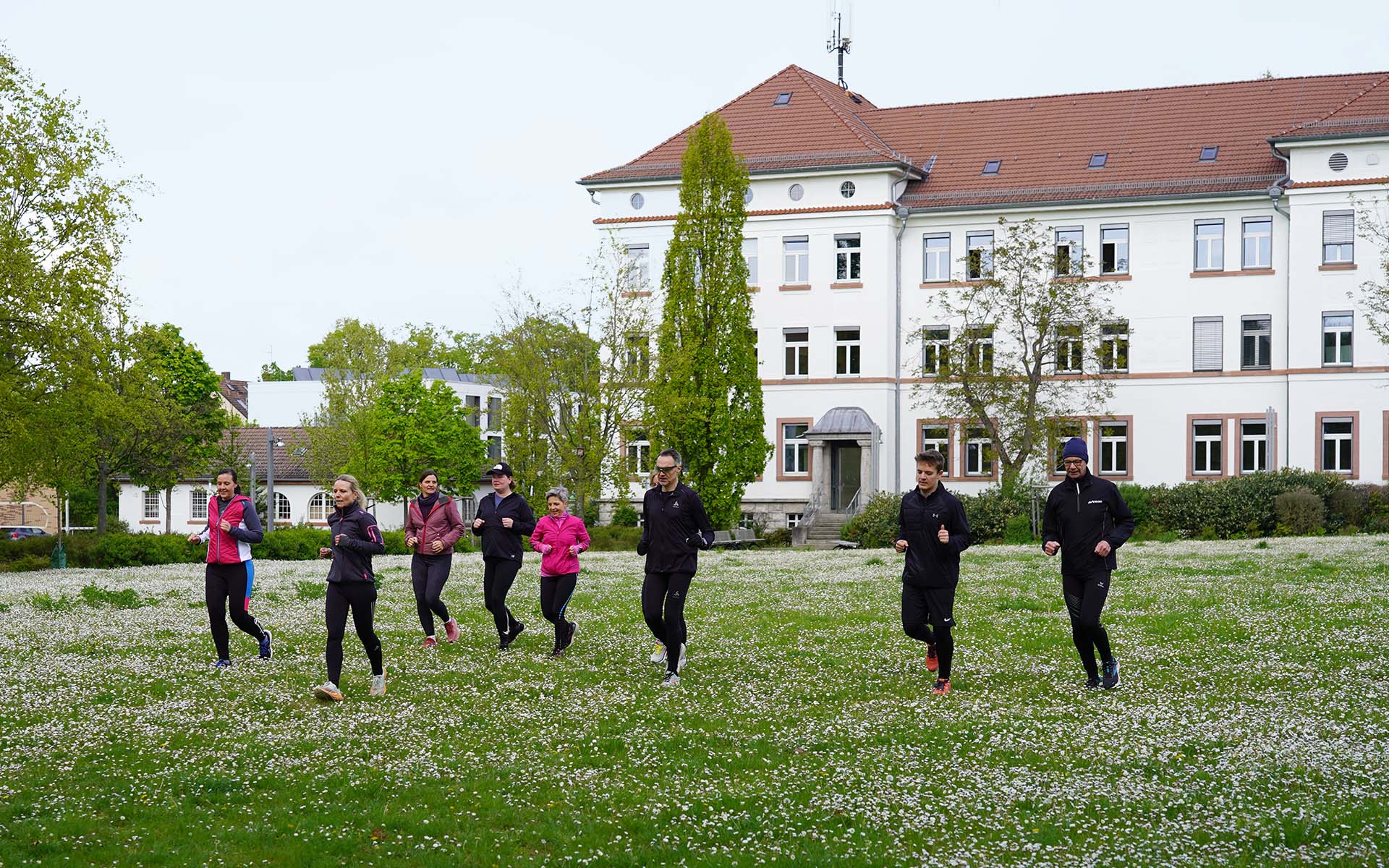 Acht Personen laufen über die grüne Campuswiese mit dem weißen Hochschulgebäude mit rotem Dach im Hintergrund