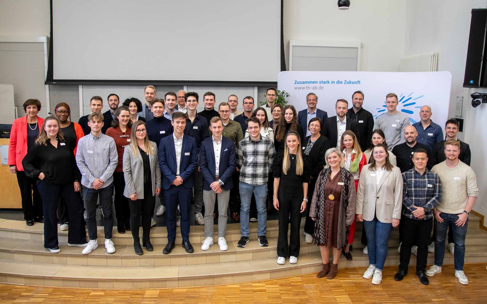 Die Stipendiatinnen und Stipendiaten des Deutschlandstipendiums zusammen mit ihren Fördernden vor der TH-Logo-Wand in der Aula