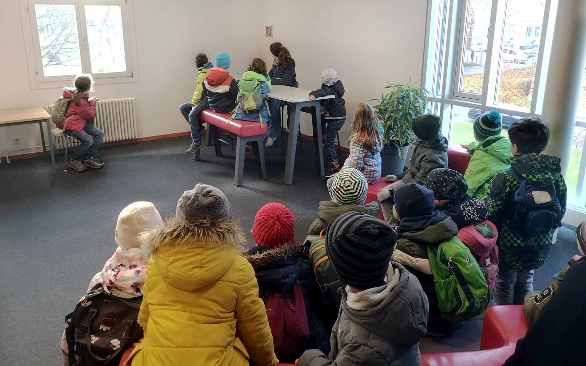Kinder sitzen gemeinsam in einem Raum der Hochschule mit dem Rücken zur Kamera