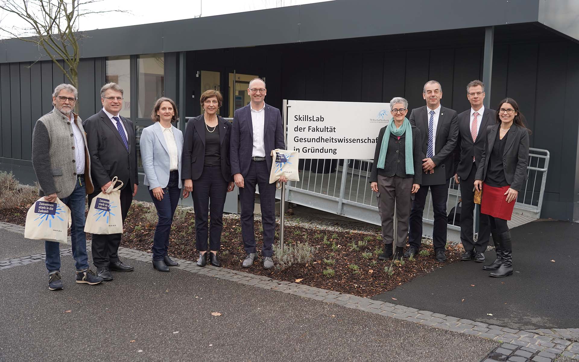Gruppenbild mit 9 Personen vor dem Schild und dem Gebäude der Fakultät Gesundheitswissenschaften