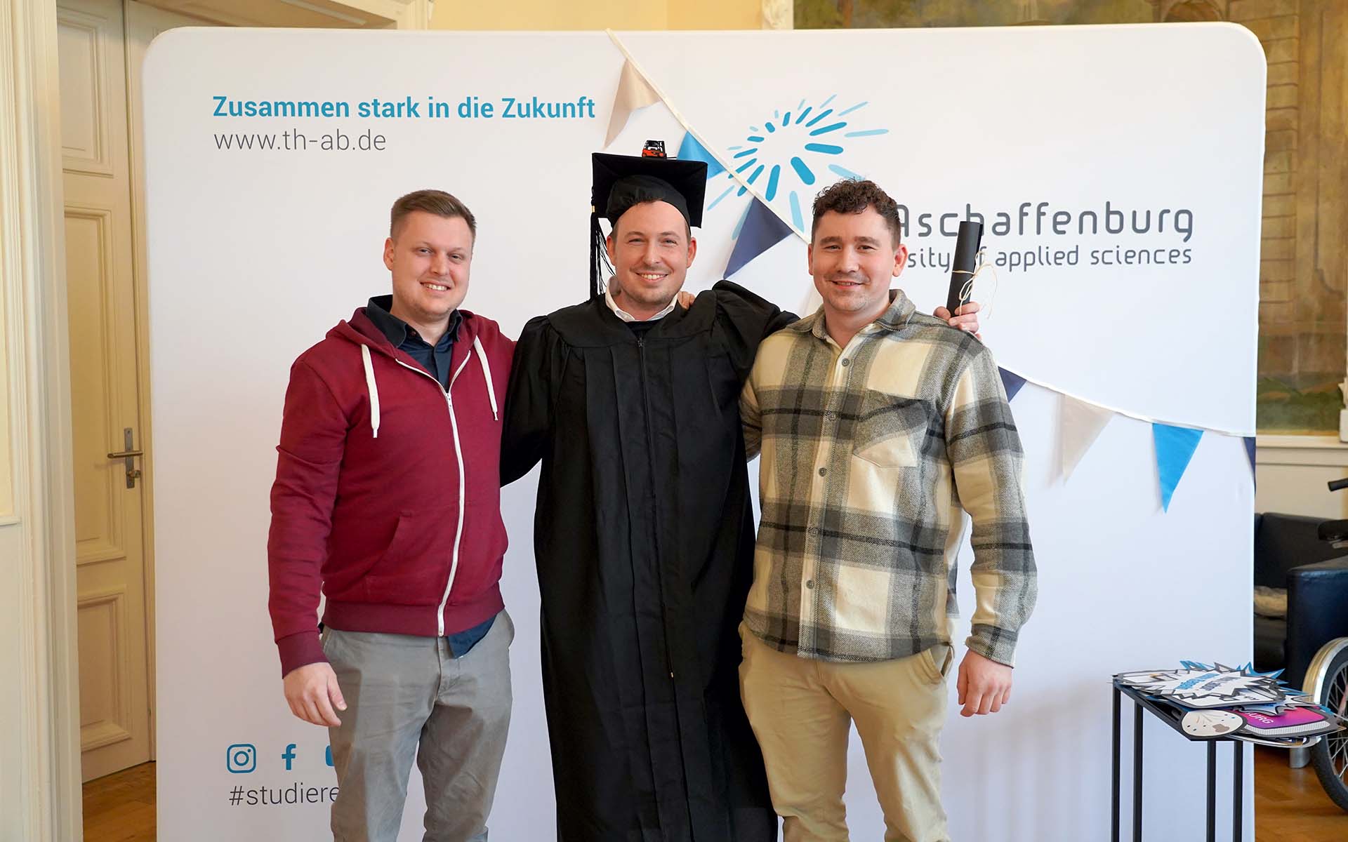 Luca Spielmann, Timm Sauer mit schwarzem Doktorhut und Umhang, Manuel Gorks Arm in Arm vor TH-Logo-Wand