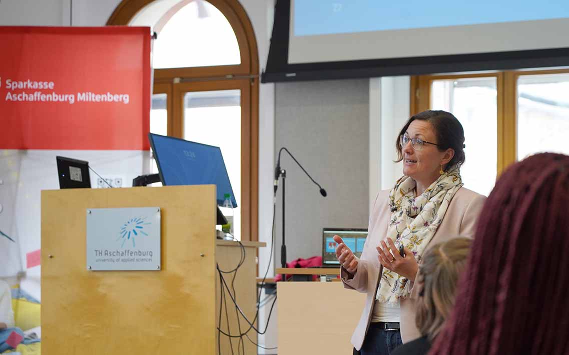 Professorin Bertels spricht am Rednerpult mit TH-Logo in der Aula, rotes Sparkassen-Roll-up dahinter