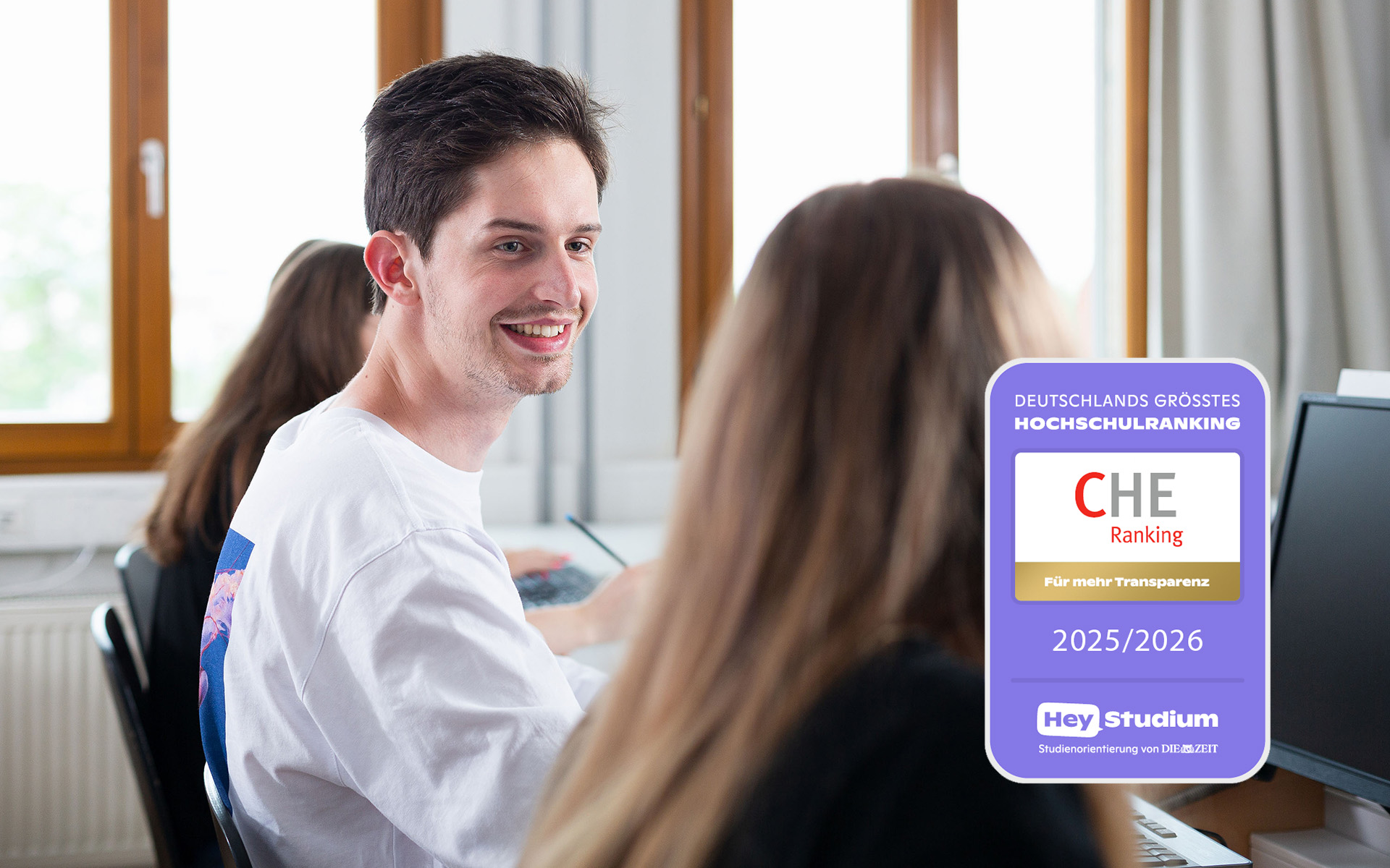 Student sitzt zwischen zwei Studentinnen im Hörsaal, CHE-Siegel auf rechter Bildhälfte