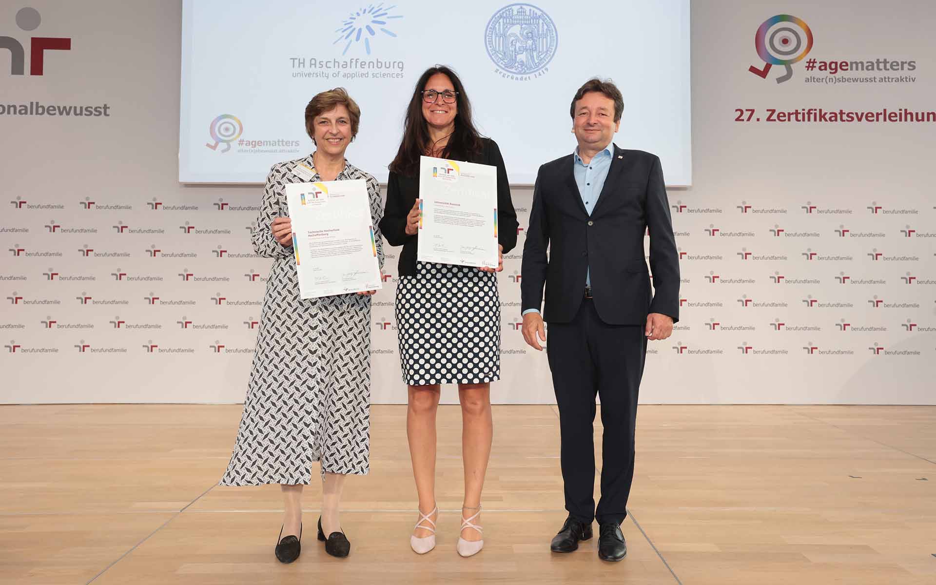 Zwei Hochschulvertreterinnen stehen mit Zertifikat in der Hand neben dem Geschäftsführer vor Logowand