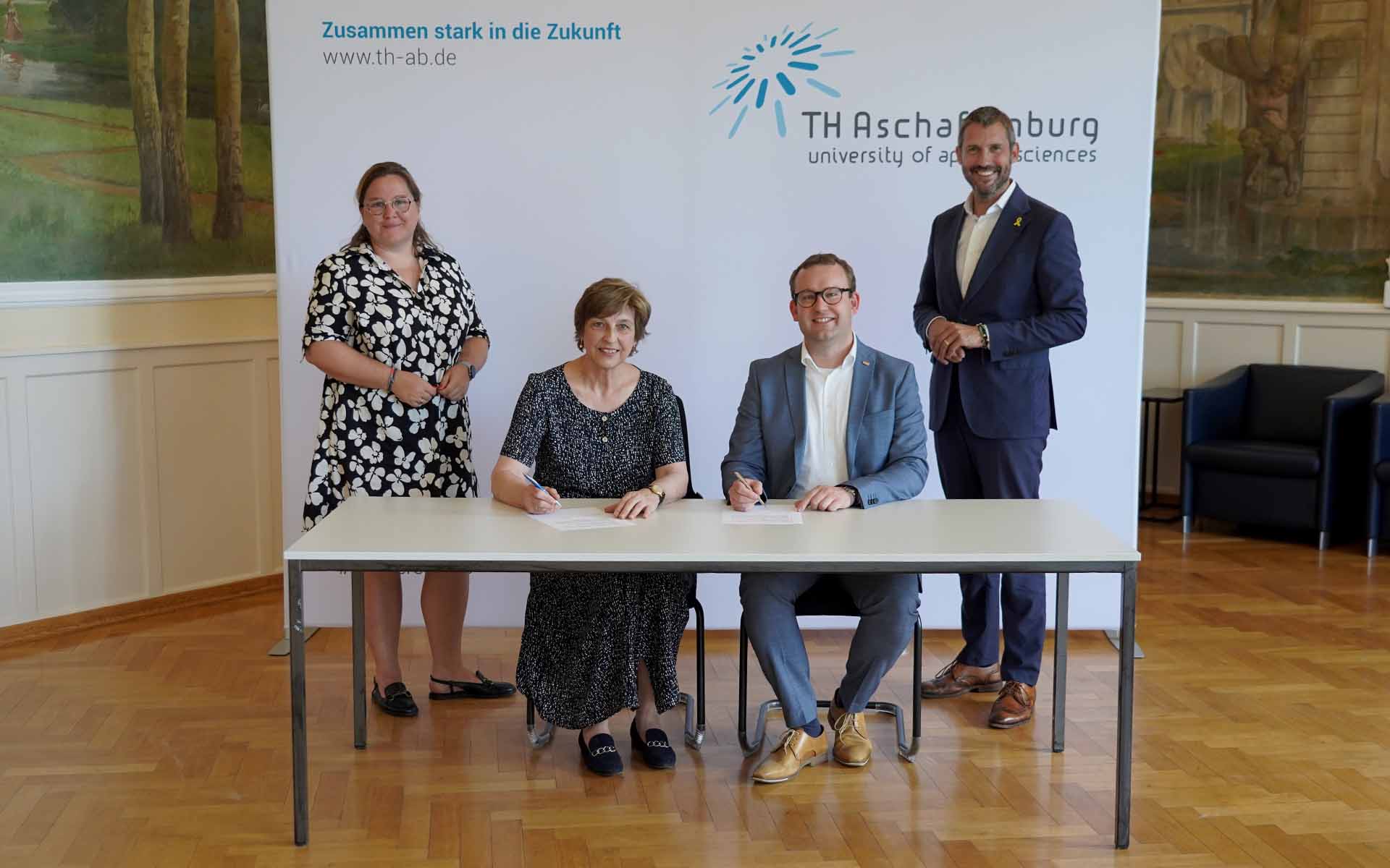 Präsidentin und BRK-Geschäftsführer sitzend bei der Vertragsunterzeichnung vor weißer Wand mit TH-Logo, Dekanin links und Landrat stehend rechts daneben