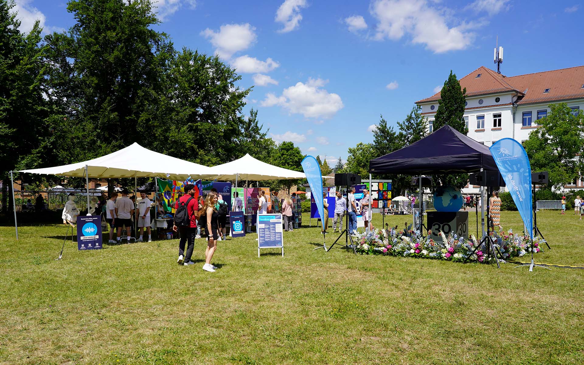 Campuswiese mit Menschen an Infoständen und weißen Zelten im Hintergrund sowie 30-Jahre-Keyvisual auf blau überdachter Bühne