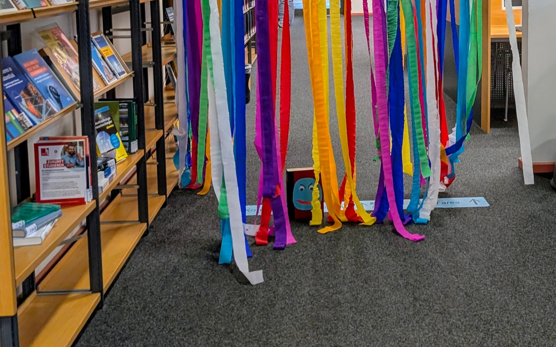 ein gebastelter Bücherwurm mit vielen bunten Papierstreifen auf dem Boden vor dem Bücherregal