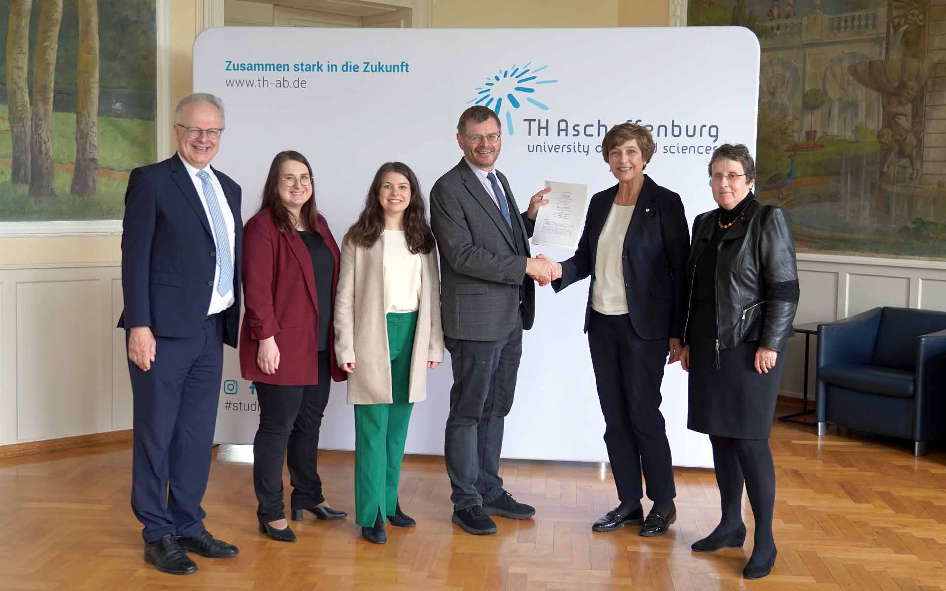 Gruppenbild mit zwei Männern und vier Frauen vor der TH-Logo-Wand im Hocksaal, Schulleiter und Präsidentin geben sich die Hand und halten den Vertrag
