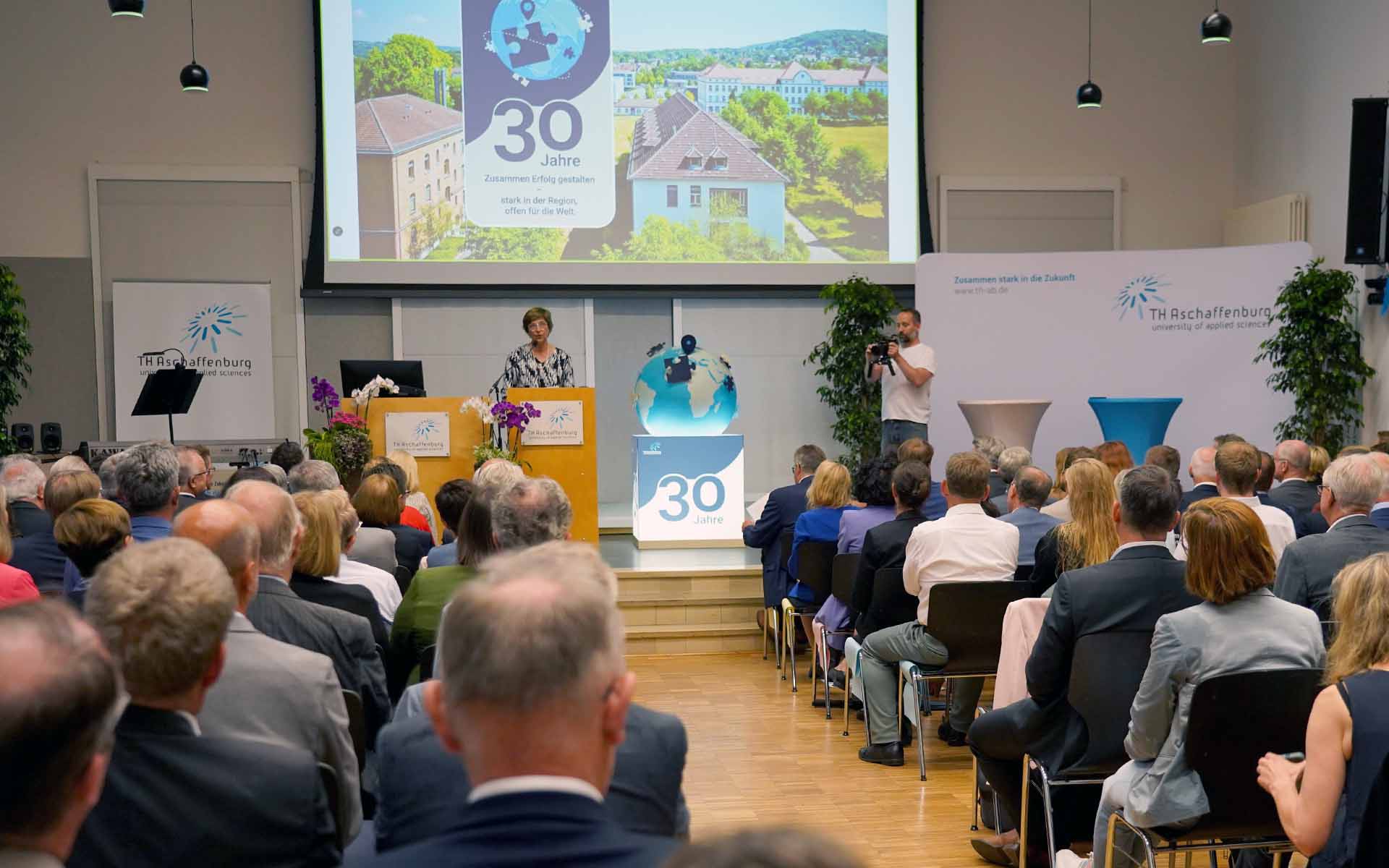 Präsidentin mit Keyvisual im Hintergrund und als Plastik auf dem Boden neben Rednerpult vor Publikum in der Aula