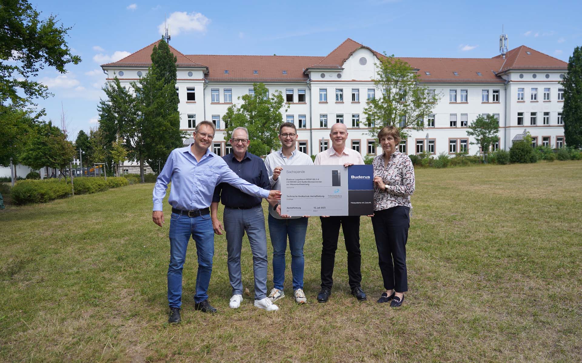 Zwei Professoren, zwei Mitarbeiter von Buderus und die Präsidentin halten auf der Campuswiese vor dem weißen Gebäude eine graue Tafel mit Firmenlogo und Bezeichnung der Wärmepumpe hoch