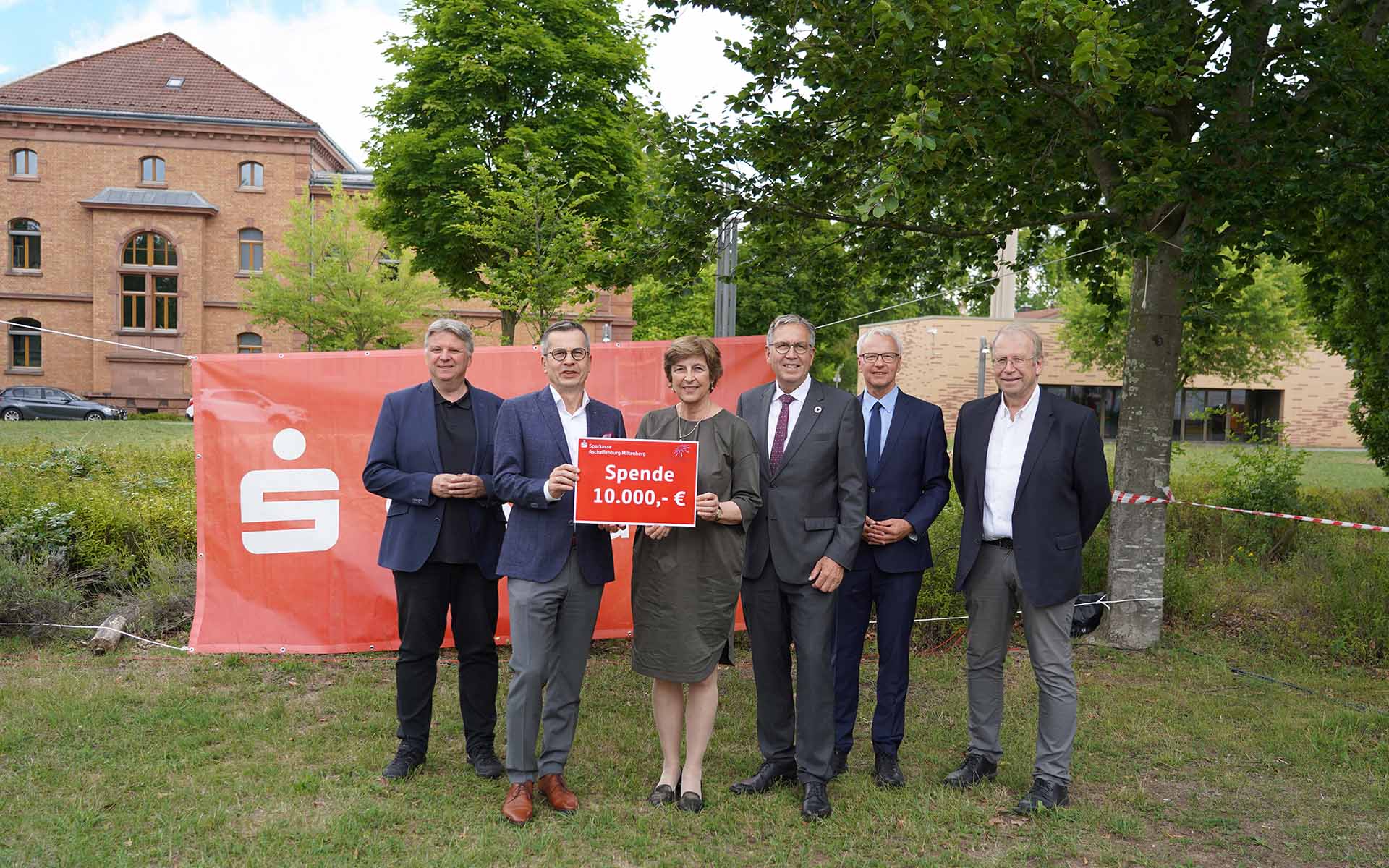 Präsidentin und Sparkassenvorstand mit rotem Schild mit Spendensumme in der Hand sowie Oberbürgermeister und drei Professoren in dunklen Anzügen stehen nebeneinander auf Campuswiese vor rotem Sparkassenbanner mit Backsteingebäude und Bäumen im Hintergrund