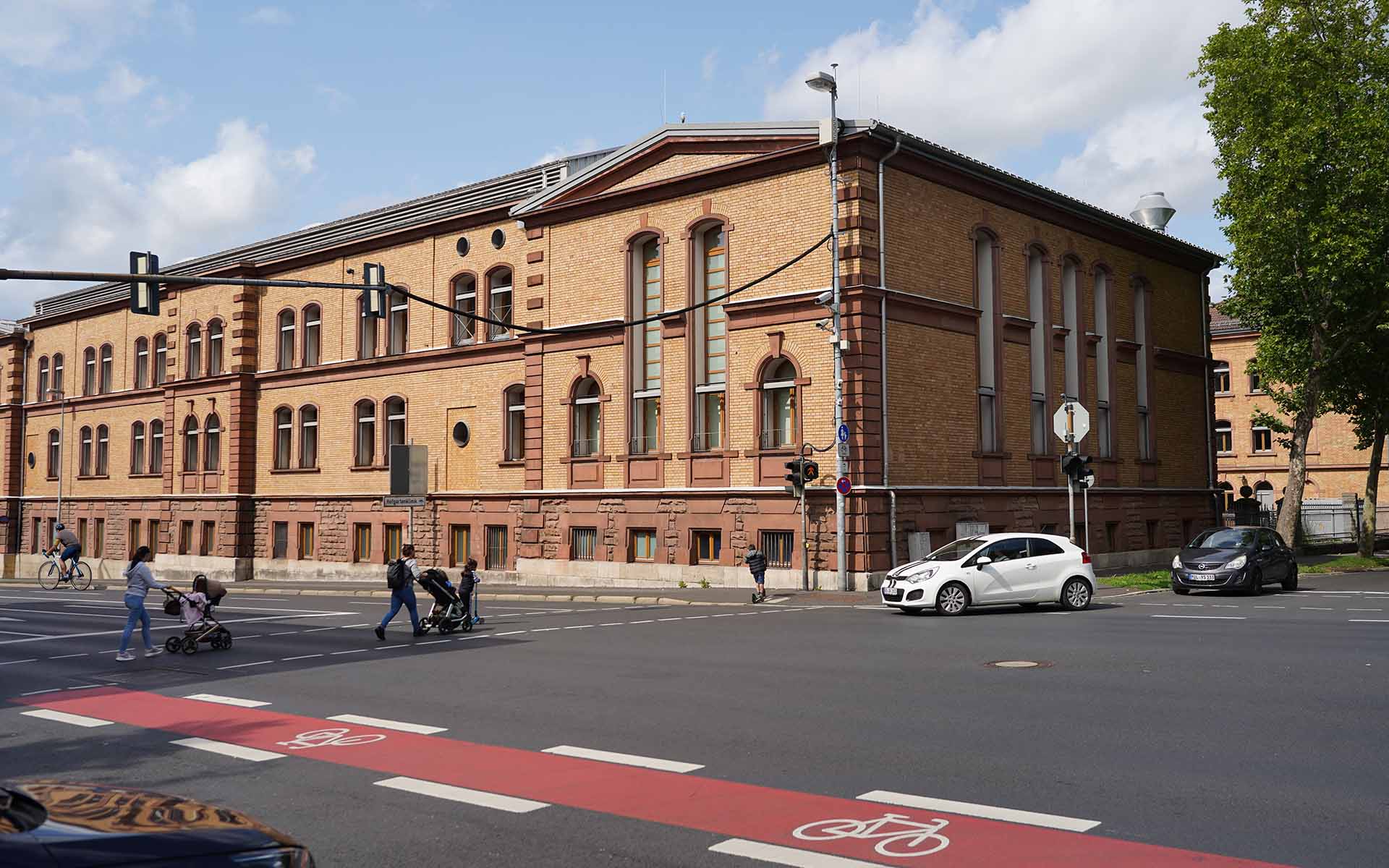 Zwei Personen mit Kinderwagen, zwei Kinder mit Rollern, ein Fahrradfahrer und zwei Autos an Ampelkreuzung vor braunem Backsteingebäude der TH