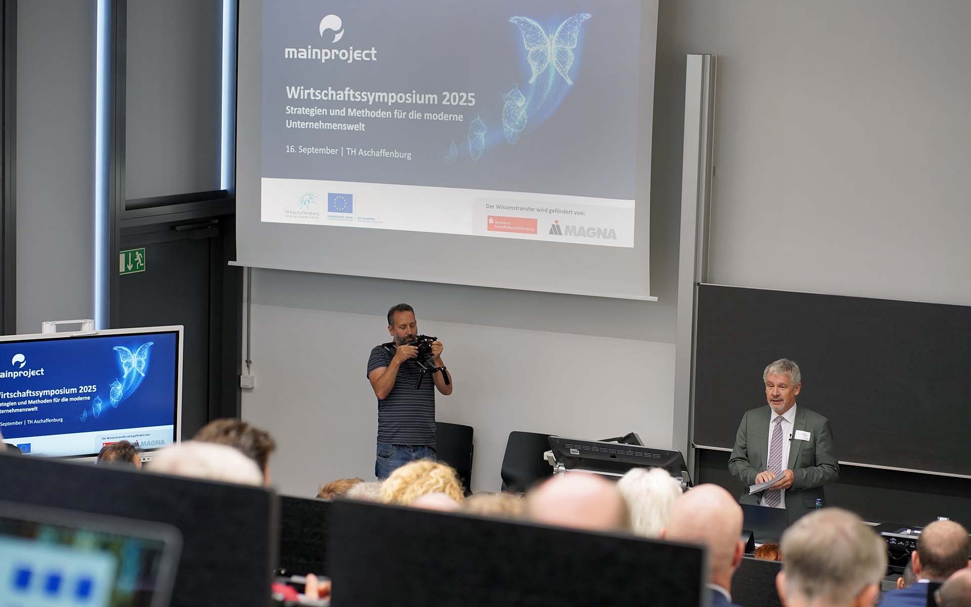 Professor Hofmann spricht vor Teilnehmenden im Hörsaal mit Beamerbild zum Wirtschaftssymposium im Hintergrund.