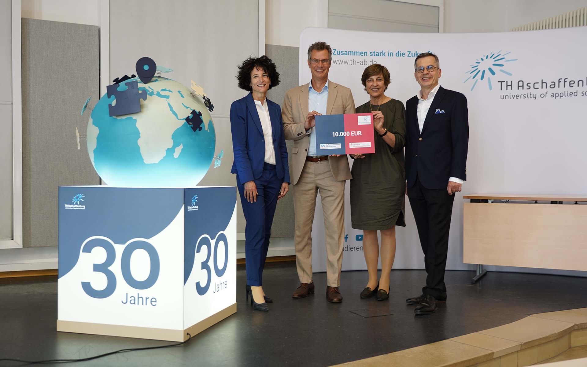 Frau in blauem Anzug, Mann mit Brille in beigem Anzug, Frau in grauem Kleid und Mann mit Brille in dunklem Anzug stehen neben beleuchteter hellblauer Weltkugel mit Würfel mit blauer 30 darauf auf Bühne der Aula mit rot-blauer Spendentafel in den Händen