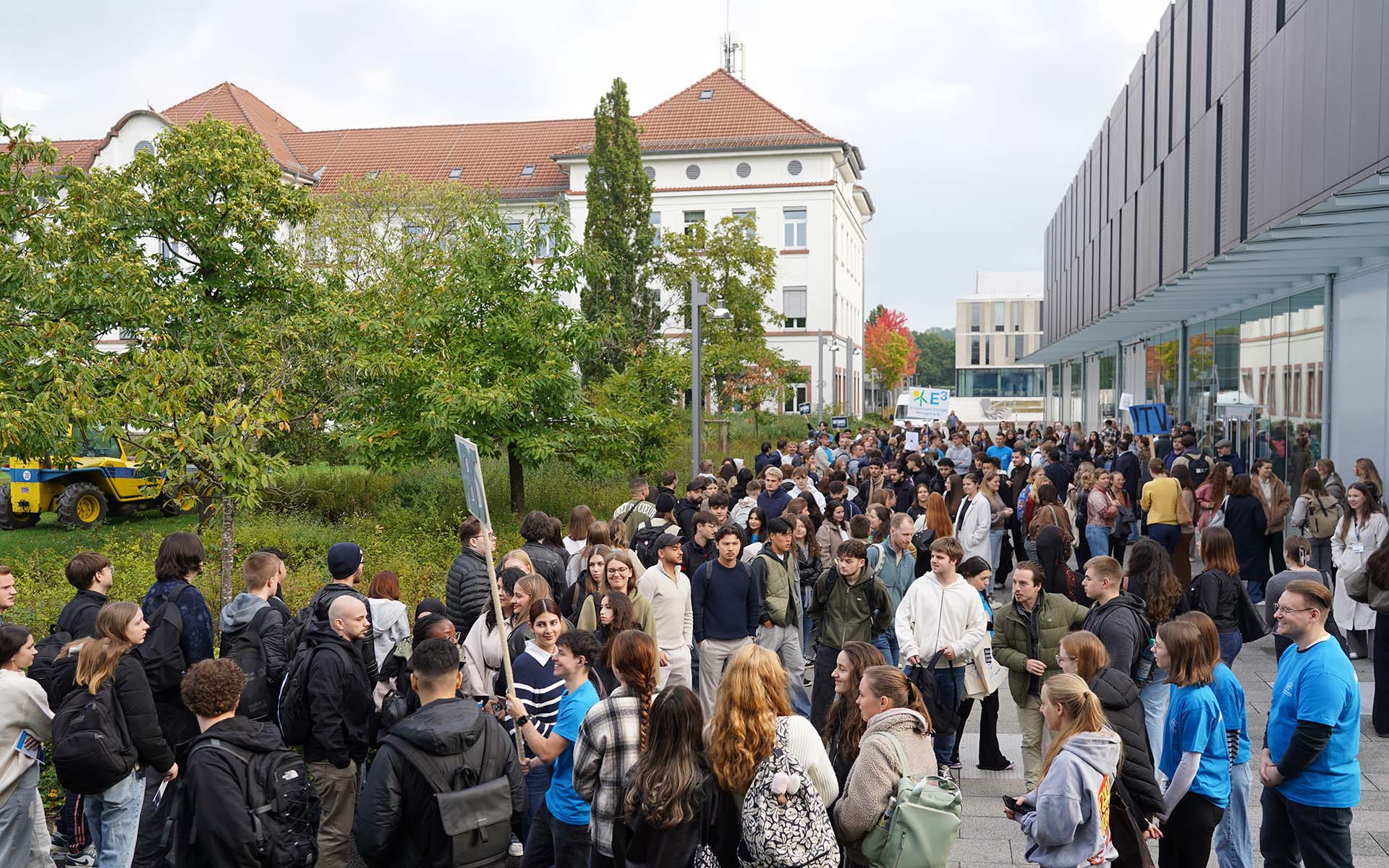 Große Menge an Studierenden zwischen einem modernen grauen Gebäude rechts und einem älteren weißen Gebäude mit rotem Dach und grünen Bäumen davor