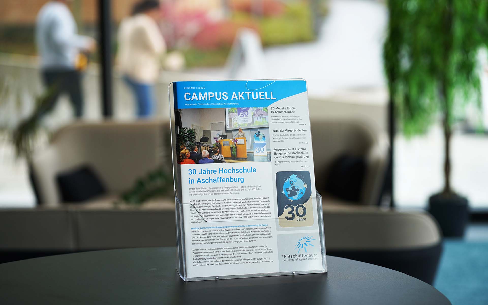 Hochschulmagazin im Prospektständer auf schwarzem Tisch im Foyer mit Sofa, Pflanze und vorbeilaufenden Personen