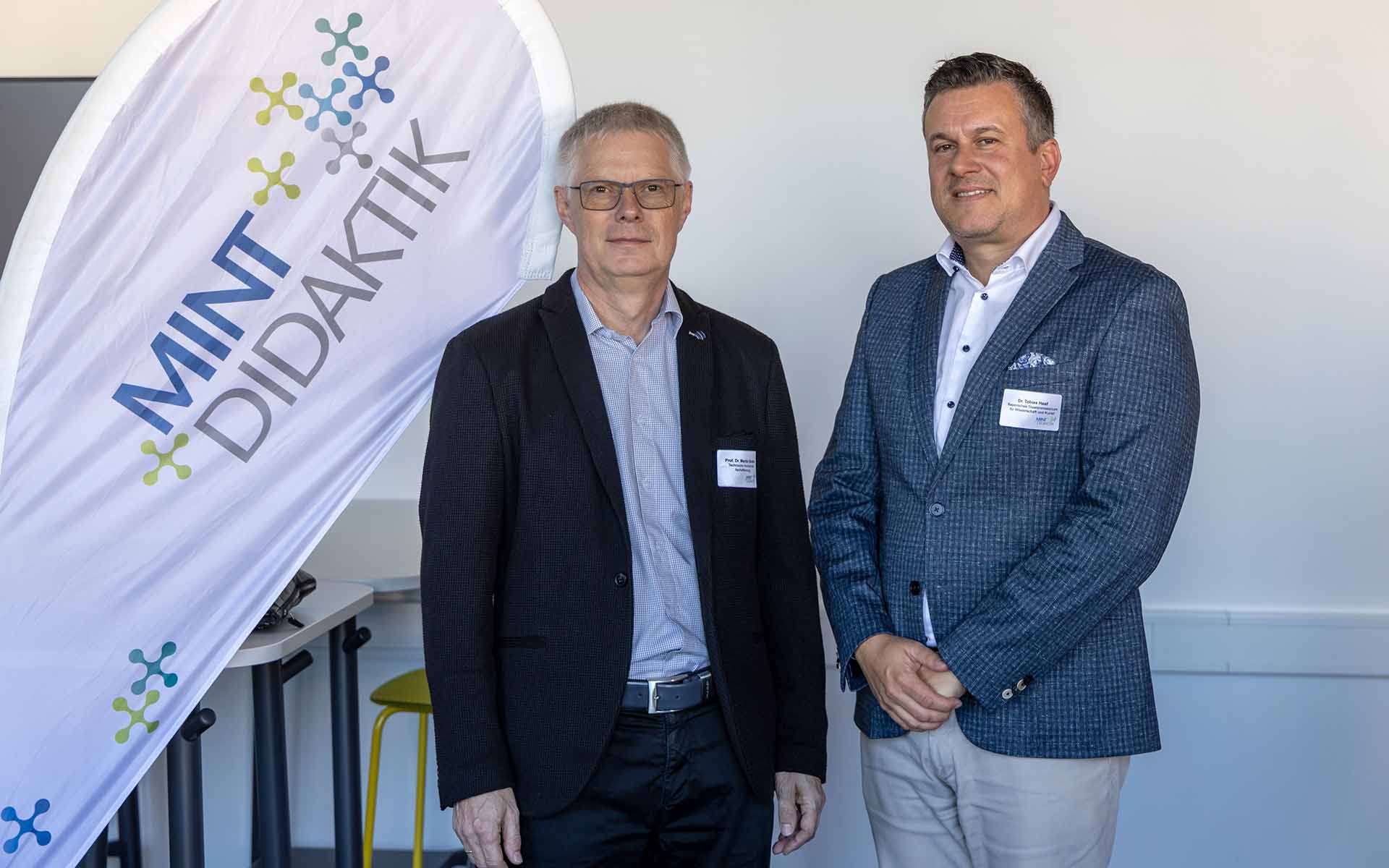 Weiße Beachflag links mit blau-grauer Aufschrift "MINT-DIDAKTIK", Mann mit grauen Haaren, Brille und dunklem Anzug Mann mit braunen Haaren, blauem Jackett und hellgrauer Hose