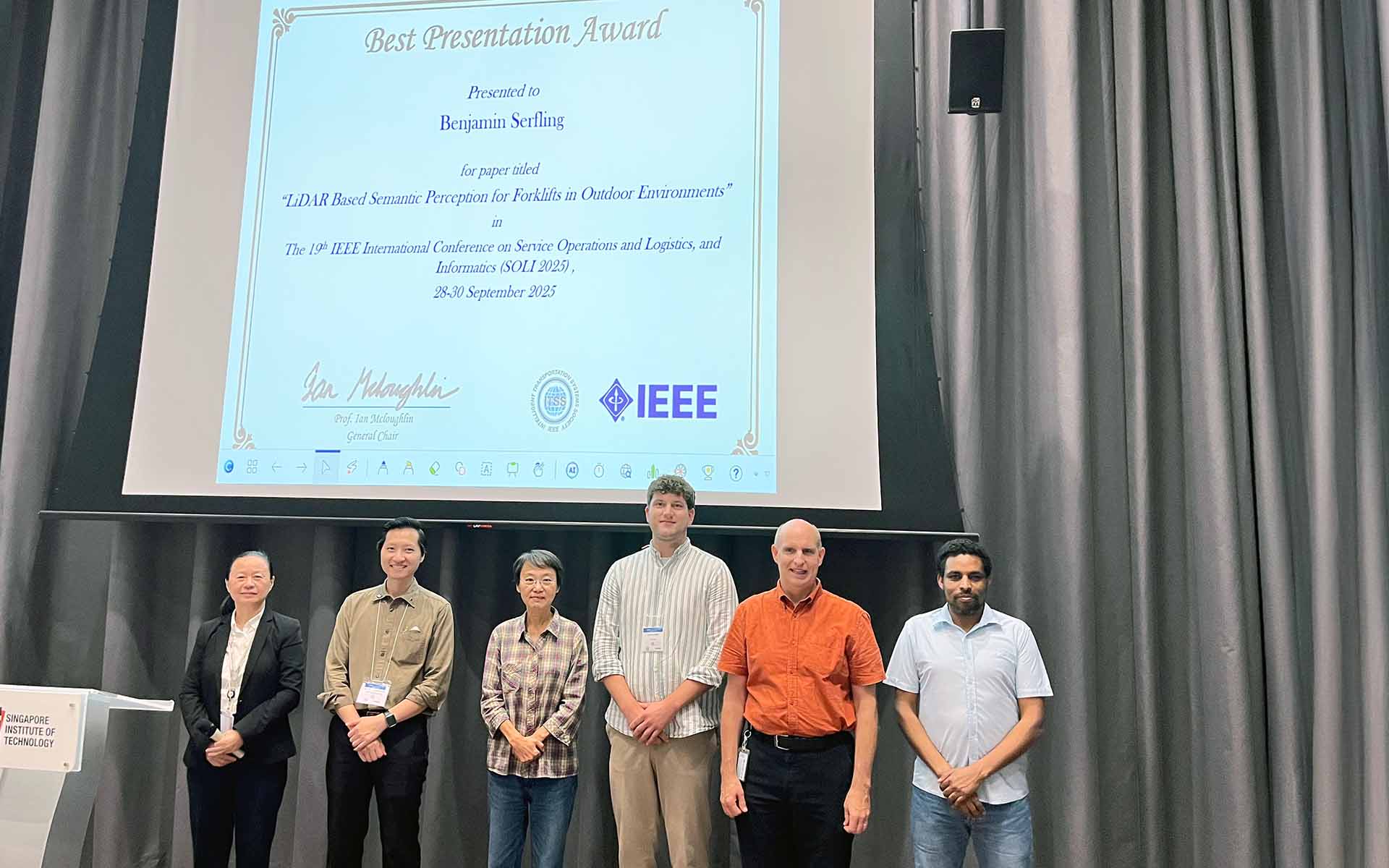 Gruppenbild mit sechs Männern und Frauen. Sie stehen vor einer Leinwand, auf der ein Bild der Urkunde vom Best Presentation Award von Benjamin Serfling abgebildet ist. 