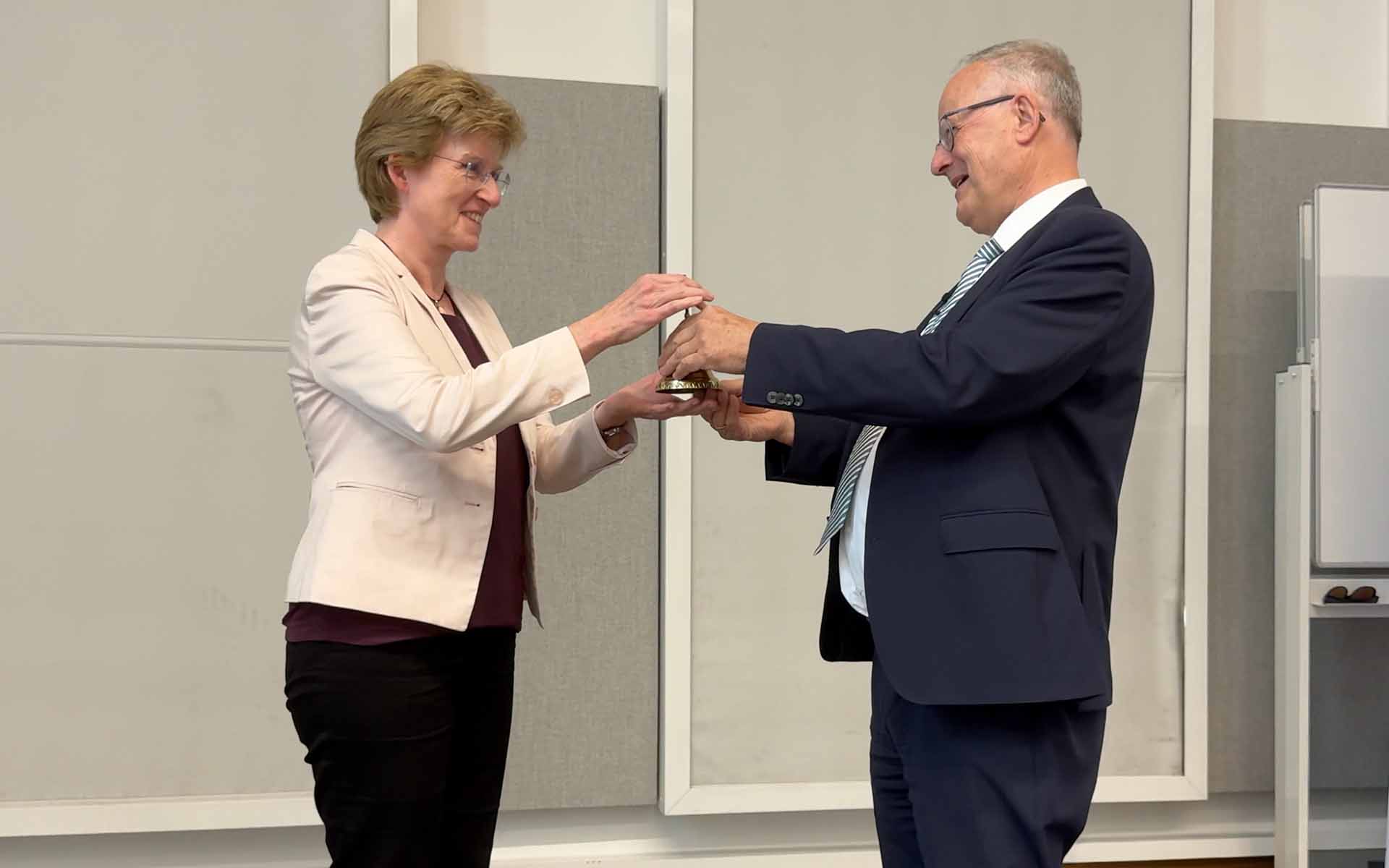 Mann mit grauen Haaren und Brille übergibt lächelnd Glocke an Frau mit kurzen blonden Haaren und Brille