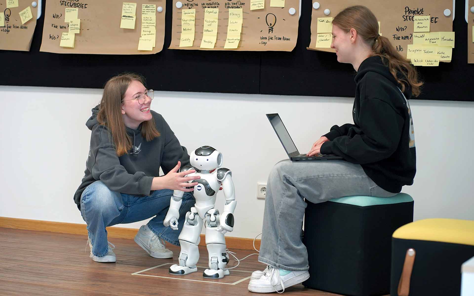 Ein Mädchen hockt neben Roboter, eines sitzt auf Hocker mit Laptop in der Hand