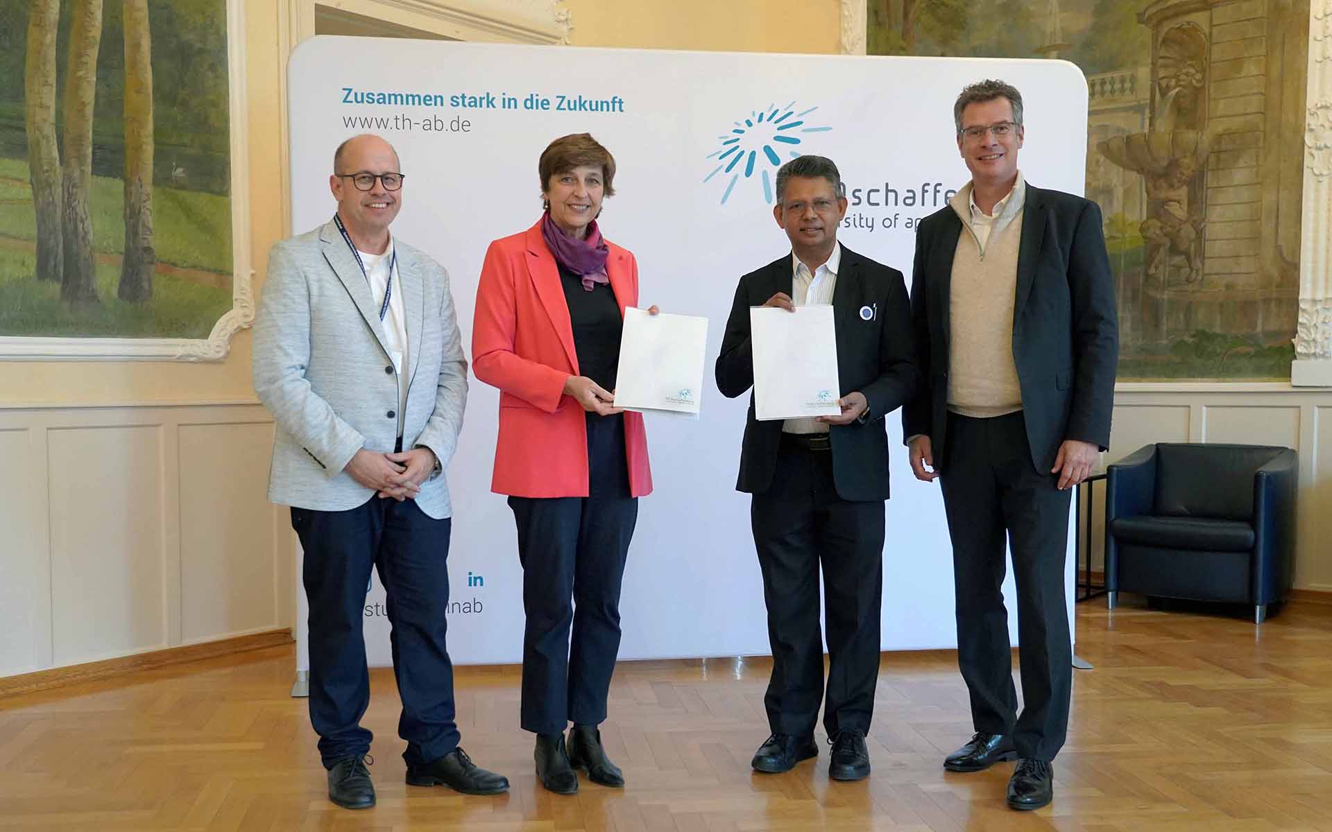 Frau mit rotem Blazer und Mann im schwarzen Anzug stehen mit Urkunden zwischen zwei Männern vor TH-Logowand