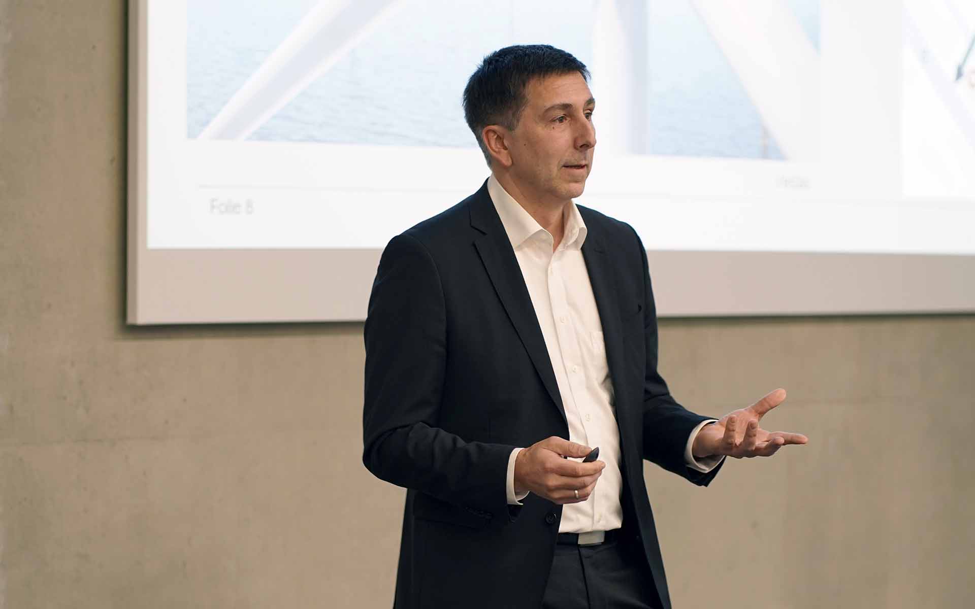 Mann mit kurzen dunklen Haaren im schwarzen Anzug spricht gestikulierend vor Leinwand