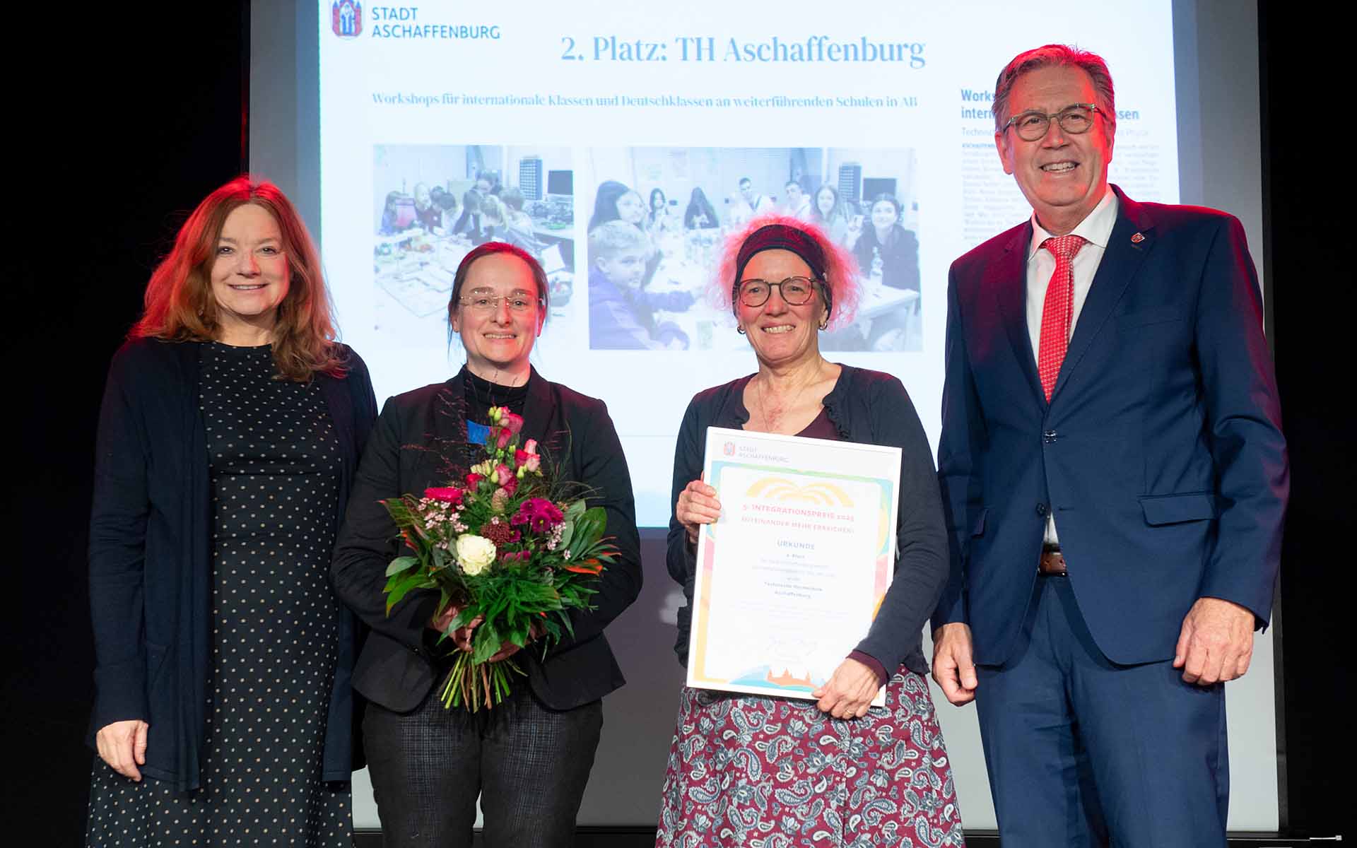 Gruppenfoto von der Urkundenverleihung mit drei Frauen, eine davon mit Urkunde, eine mit Blumenstrauß und einem Mann
