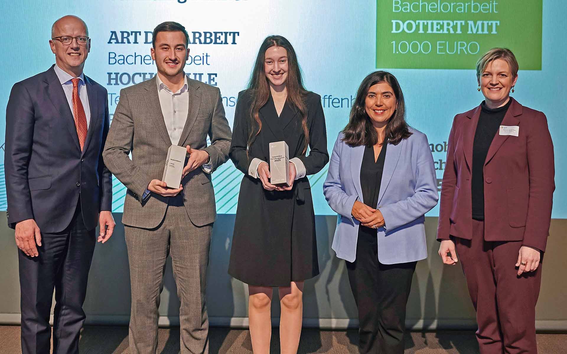 Fünf Menschen stehen vor einer Leinwand. Zwei von ihnen halten Preise in der Hand