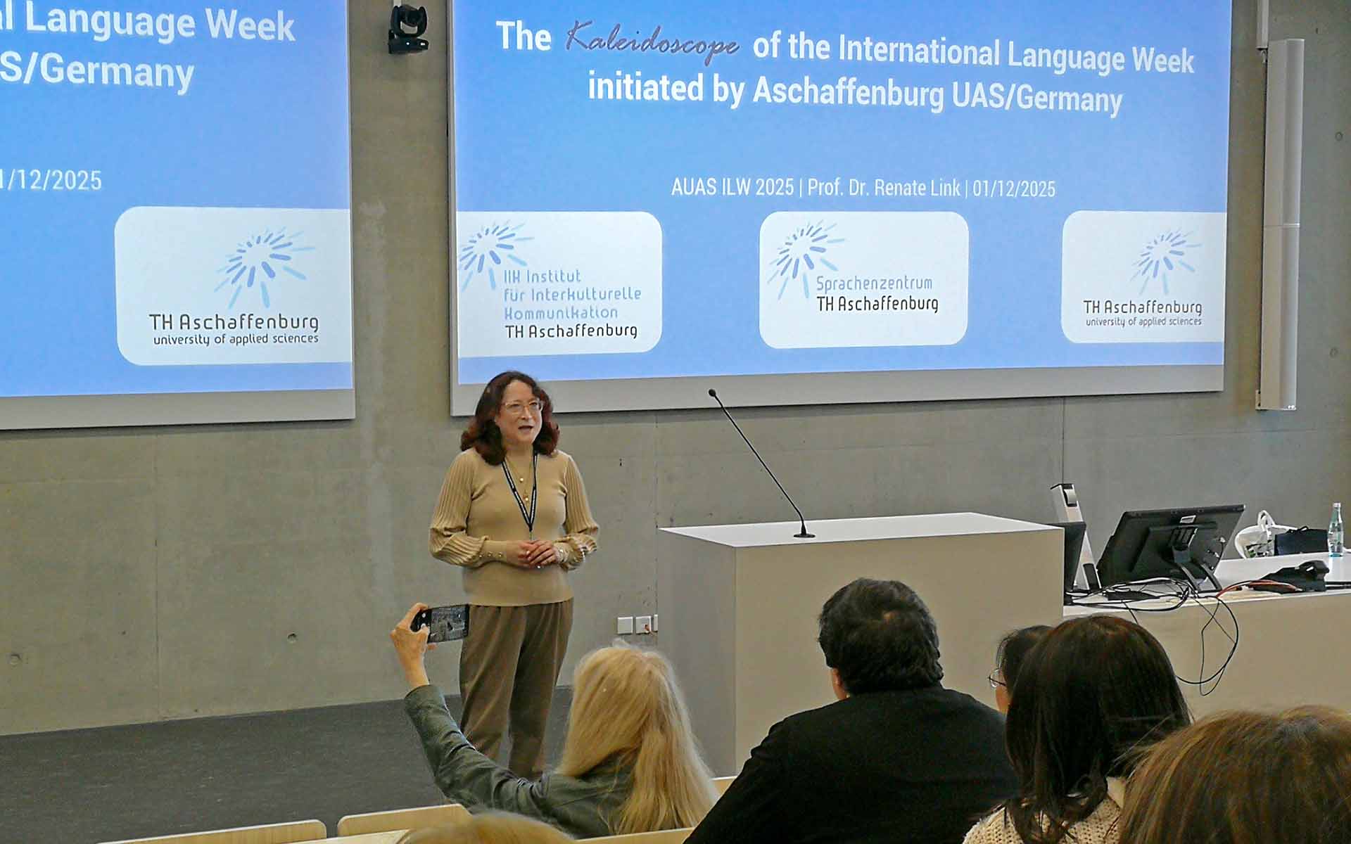 Frau mit roten schulterlangen Haaren und Brille spricht vor cyanblauer Präsentation zum Publikum