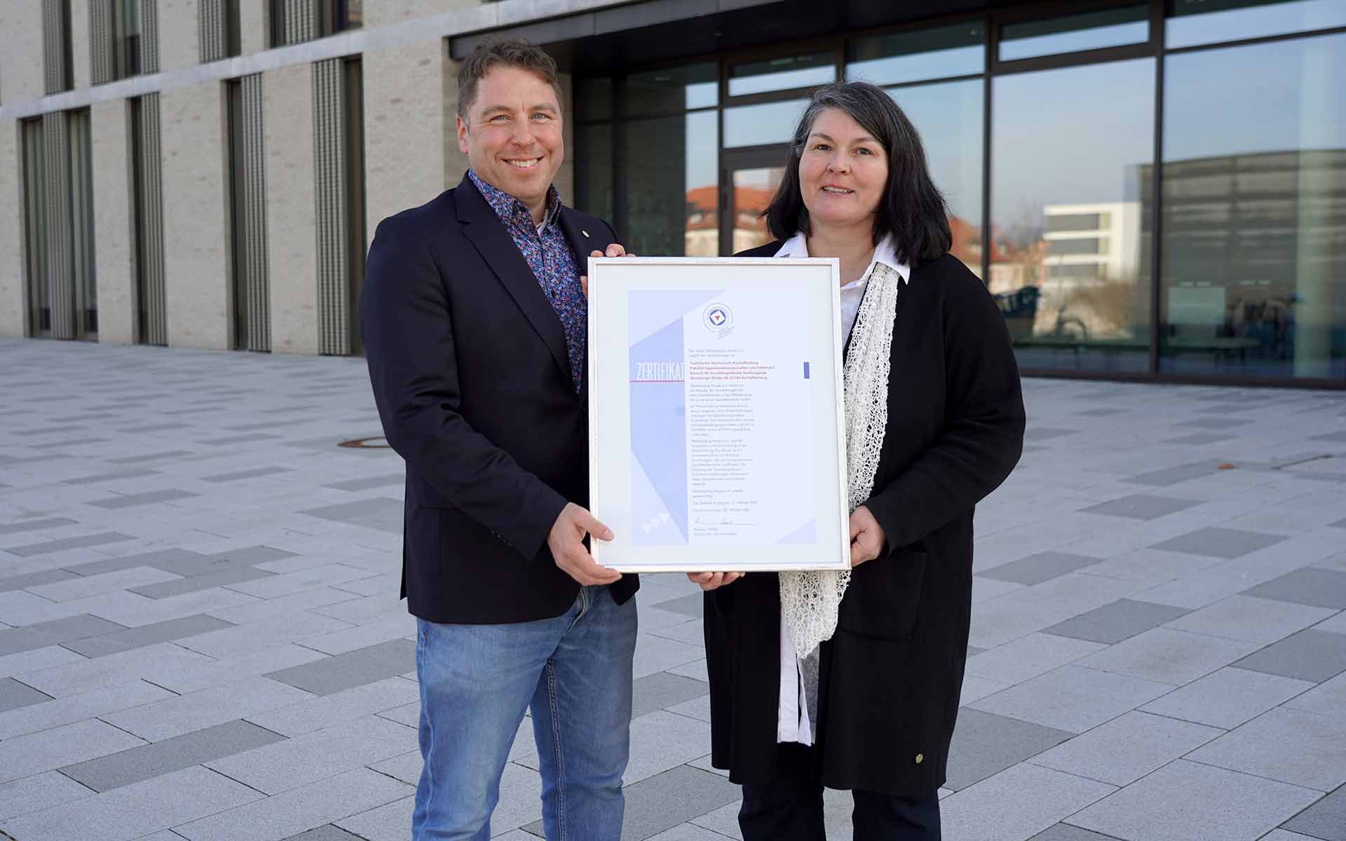 Ein Mann und eine Frau stehen vor einem TH-Gebäude mit der Urkunde in der Hand