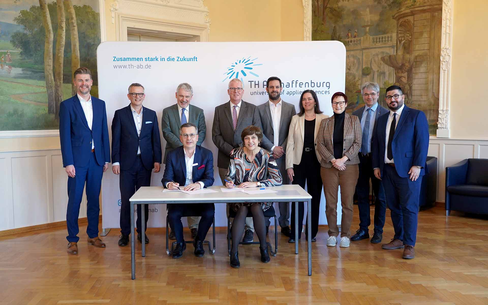 Zwei sitzende Personen, die Vertrag unterzeichnen und neun Personen stehen dahinter