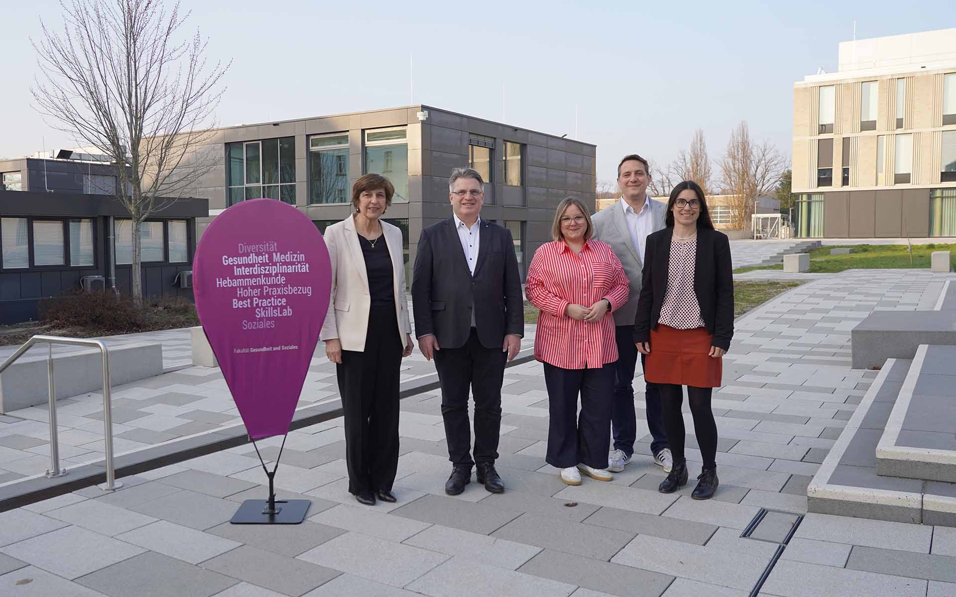 Drei Frauen und zwei Männer stehen vor dem Skills-Lab und dem Neubau neben einer lilafarbenen Pinnflag mit Begriffen der Fakultät GS