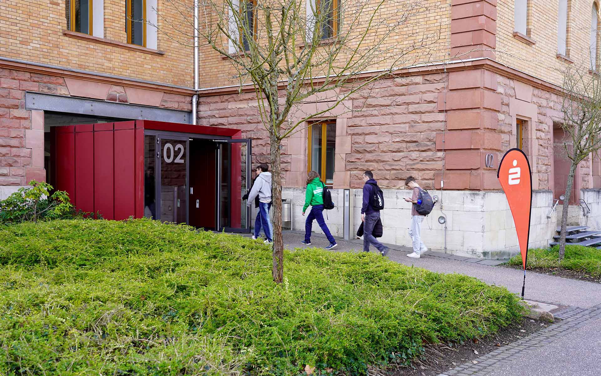 Vier Schülerinnen und Schüler laufen auf Eingangstür von TH-Gebäude an Sparkassen-Dropflag vorbei