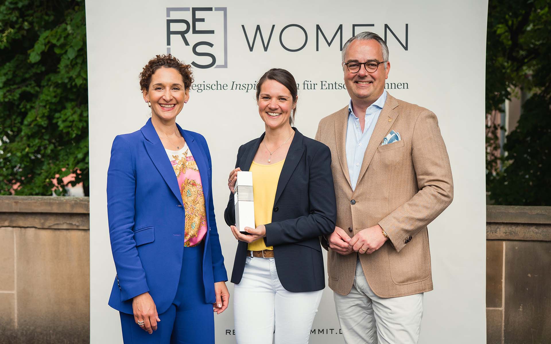 Eine lächelnde Frau steht mit einem weißen, säulenartigen Gegenstand in der Hand zwischen einem lächelnden Mann und einer lächelnden Frau vor einer weißen Wand mit schwarzer Schrift