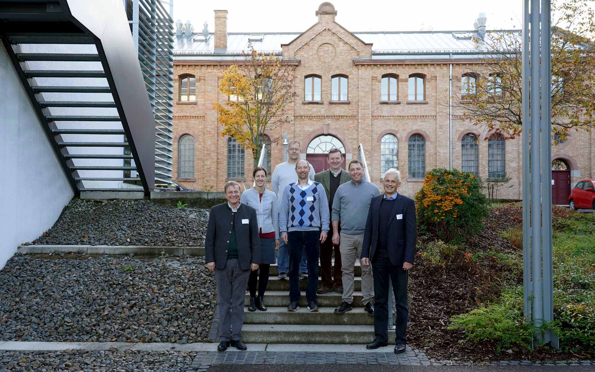 Sechs Männer und eine Frau stehen auf einer Treppe vor einem Gebäude mit einer rotbraunen Fassade