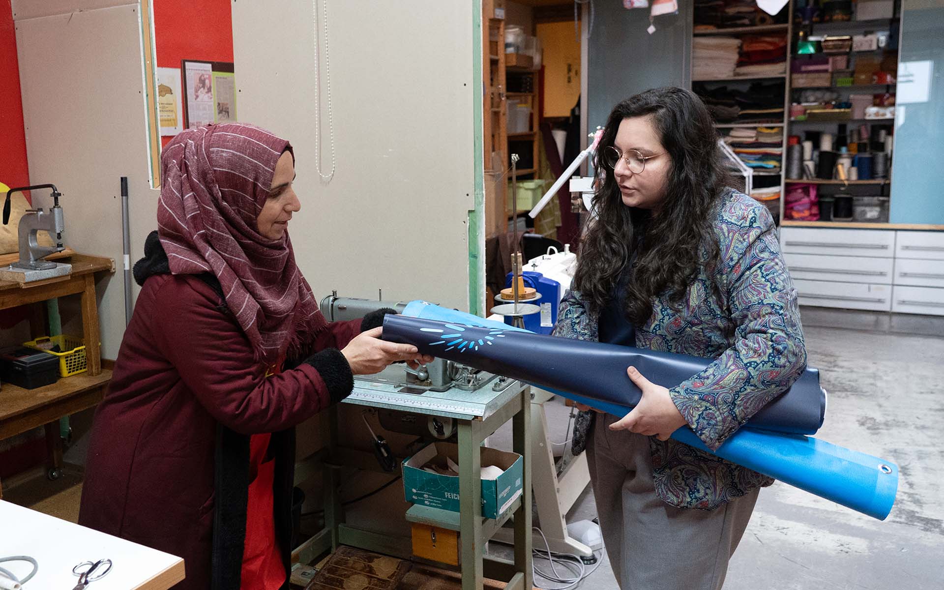 In der Nähwerkstatt übergibt Alexandra Koch Amina von Gesta e.V. alte, blaue Werbebanner der TH Aschaffenburg