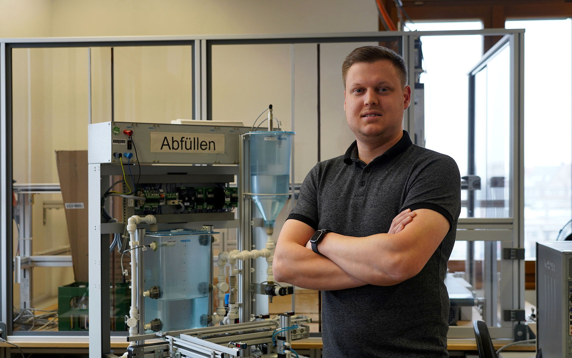 Luca Spielmann steht mit verschränkten Armen und in die Kamera lächelnd vor einer Maschine die mit dem Wort "Abfüllen" beschriftet ist.