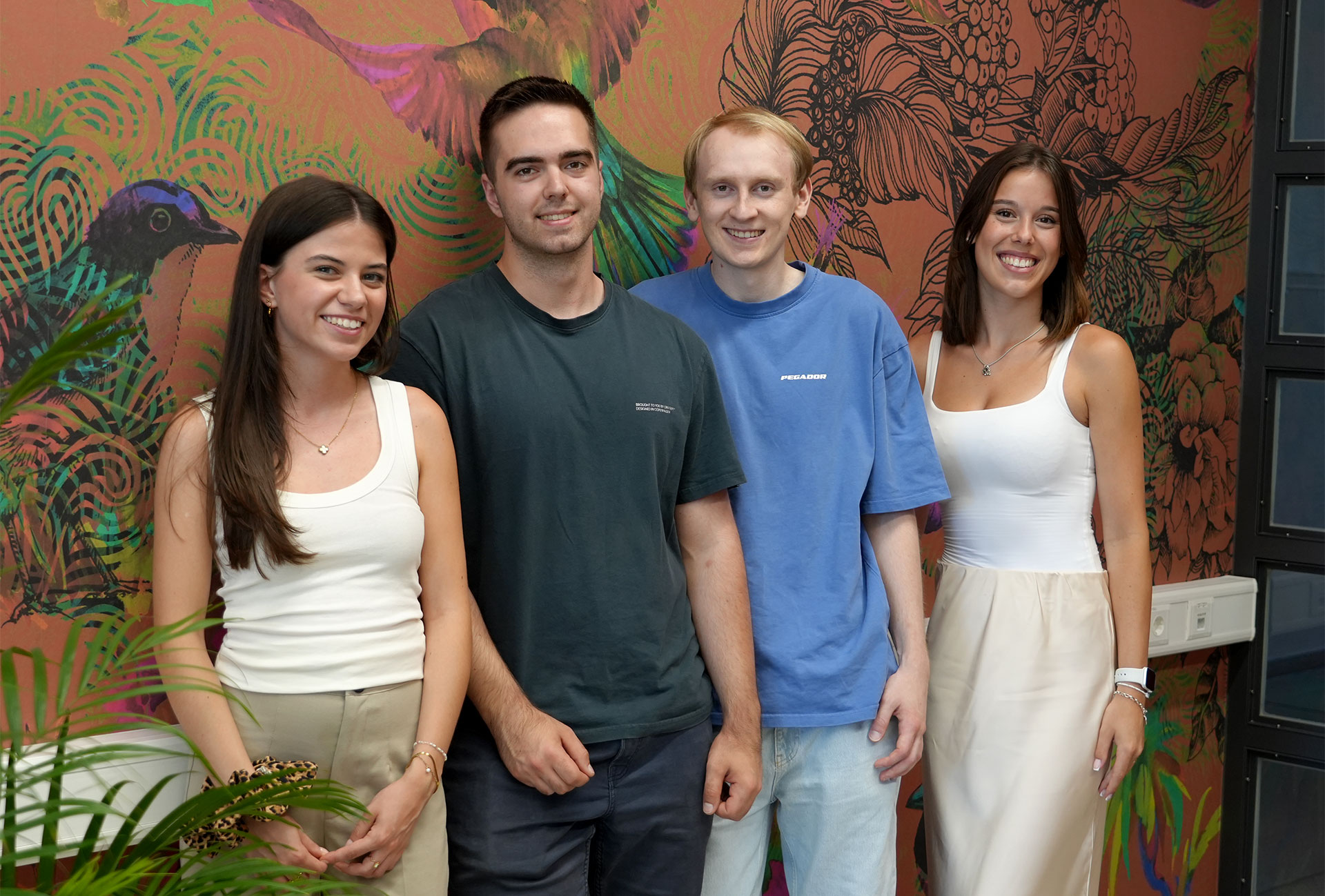 Vier Studierende, Christoph, Clarissa, Emilia und Marc, stehen lächelnd vor einer bunten Wand